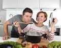 Young attractive couple at home kitchen with man tasting vegetable stew cooked by her wife smiling happy