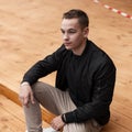 Young attractive fashionable man in a vintage stylish black jacket in beige trousers relaxes sitting on wooden steps outdoors Royalty Free Stock Photo