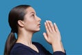 Young female pleading, holding hands in prayer on blue background, profile view