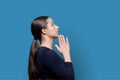 Young female pleading, holding hands in prayer on blue background, profile view
