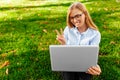 Young attractive girl in glasses, sitting with a laptop, talking on Skype and showing two fingers, on a laptop screen, in a park o Royalty Free Stock Photo