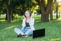 A young and attractive girl in headphones sitting on the grass on the lawn. Using laptop to listening to music with headphones. Royalty Free Stock Photo