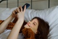 A young attractive girl is lying in bed with a smartphone, portrait, leafing through the news on the Internet and smiling Royalty Free Stock Photo