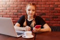 Young attractive girl surfing internet via mobile phone and smiling while sitting alone in coffee shop during working on laptop, Royalty Free Stock Photo