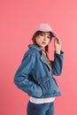 Young attractive girl with two braids in denim jacket and cap with headphones on neck holding hand in pocket dreamly