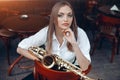 Young attractive girl in white shirt with a saxophone sitting on caffe shop - outdoor in street. young woman with sax looking Royalty Free Stock Photo