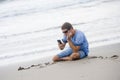Attractive and handsome man on his 30s sitting on the sand relaxed on the beach laughing in front of the sea texting on mobile pho Royalty Free Stock Photo