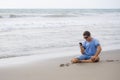 Attractive and handsome man on his 30s sitting on the sand relaxed on the beach laughing in front of the sea texting on mobile pho Royalty Free Stock Photo