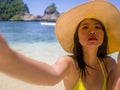 Young attractive and happy Asian Chinese woman in pamela Summer hat and yellow bikini taking selfie with phone at beautiful beach Royalty Free Stock Photo