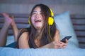 Young attractive and happy Asian Chinese woman with yellow headphones listening to music in mobile phone on bed at home smiling ha Royalty Free Stock Photo