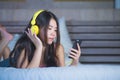 Young attractive and happy Asian Chinese woman with yellow headphones listening to music in mobile phone on bed at home smiling ha Royalty Free Stock Photo