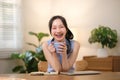 Young attractive happy Asian female student holding a cup of coffee sitting in the living room smiling and looking at Royalty Free Stock Photo