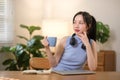 Young attractive happy Asian female student holding a cup of coffee sitting in the living room smiling and looking at Royalty Free Stock Photo