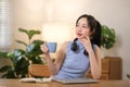 Young attractive happy Asian female student holding a cup of coffee sitting in the living room smiling and looking at Royalty Free Stock Photo