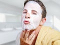 Young attractive and happy gay man in bathroom using beauty paper facial mask touching his face looking to the mirror smiling in Royalty Free Stock Photo