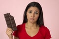 Attractive and happy hispanic woman in red top feeling guilty holding chocolate bar on pink background Royalty Free Stock Photo