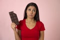 Attractive and happy hispanic woman in red top feeling guilty holding chocolate bar on pink background Royalty Free Stock Photo