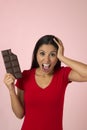 Young attractive and happy hispanic woman in red top smiling excited eating chocolate bar on pink background