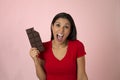 Young attractive and happy hispanic woman in red top smiling excited eating chocolate bar on pink background