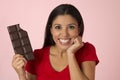 Young attractive and happy hispanic woman in red top smiling excited eating chocolate bar on pink background