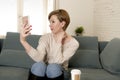 young attractive and happy red hair woman sitting at home sofa couch drinking coffee taking selfie picture with mobile phone Royalty Free Stock Photo