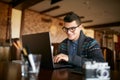 Smiling Attractive Hipster Man In Glasses And Winter Sweater Working On Laptop In Cafe. Freelancer Businessman Browsing