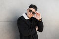 Young attractive hipster man in a trendy baseball cap in stylish sunglasses in a vintage black jacket with a white collar poses Royalty Free Stock Photo