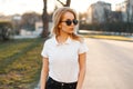 Young attractive hipster woman in stylish sunglasses in a white polo t-shirt in black jeans walking in downtown at sunset. Royalty Free Stock Photo