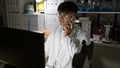 Young attractive hispanic man, a dynamic scientist, juggling technology in lab, talking on smartphone while working on computer Royalty Free Stock Photo