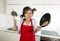 Young attractive home cook woman in red apron at kitchen holding pan and household with pot on her head in stress