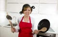 Young attractive home cook woman in red apron at kitchen holding pan and household with pot on her head in stress