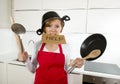 Young attractive home cook woman in red apron at kitchen holding pan and household with pot on her head in stress