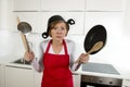 Young attractive home cook woman in red apron at kitchen holding pan and household with pot on her head in stress