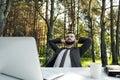 Young attractive male businessman in suit sitting at desk in forest of nature park. Stretches and rests from work. Royalty Free Stock Photo