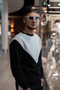 Young attractive man with a beard in an American baseball cap in white vintage sunglasses in a fashionable sweatshirt stands