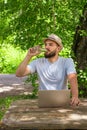 A young attractive man with a beard drinks clear water from a bottle while working at a computer. Sitting in a park.