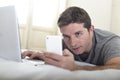 Young attractive man lying on bed or couch using mobile phone and computer laptop internet addict Royalty Free Stock Photo