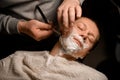 Young attractive man relaxing while sitting in barber& x27;s chair and getting shave by male barber