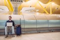 Young attractive man sitting at the airport working with a laptop waiting his flight with a suitcase Royalty Free Stock Photo