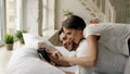 Young attractive man using tablet computer lying in bed while his girlfriend come and hug him in bedroom at the morning Royalty Free Stock Photo