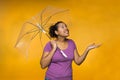 Young attractive mixed race woman holding an umbrella checking for rain wearing a purple shirt against a yellow background