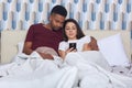 Young attractive multicultural couple in white bed, female holding smartphones in hands, wearing pajamas, searching or browsing Royalty Free Stock Photo