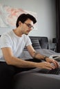 Young attractive pensive brunette man in T-shirt and eyeglasses sitting on bed and dreamily working on laptop at modern Royalty Free Stock Photo