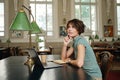 Young attractive pensive student dreamily studying on laptop in library of university