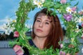 A young attractive red-haired girl looks through a wreath of wild flowers into the lens on a warm summer day.