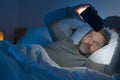 Young attractive and relaxed man with blue eyes lying on bed late at night in dark and dim light networking on mobile phone or Royalty Free Stock Photo