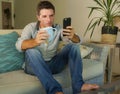 Young attractive and relaxed man at home using mobile phone drinking coffee sitting at living room sofa couch dating or flirting o Royalty Free Stock Photo