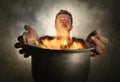 Young attractive and shocked messy home cook man with apron holding cooking pot in fire burning the food in kitchen disaster Royalty Free Stock Photo