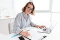 Young attractive smiling girl in glasses and striped shirt working with documents and computer while siting at table in light kit Royalty Free Stock Photo