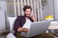 Young attractive smiling guy is browsing at his laptop and talking oh mobile phone, sitting at home on the cozy sofa, wearing Royalty Free Stock Photo